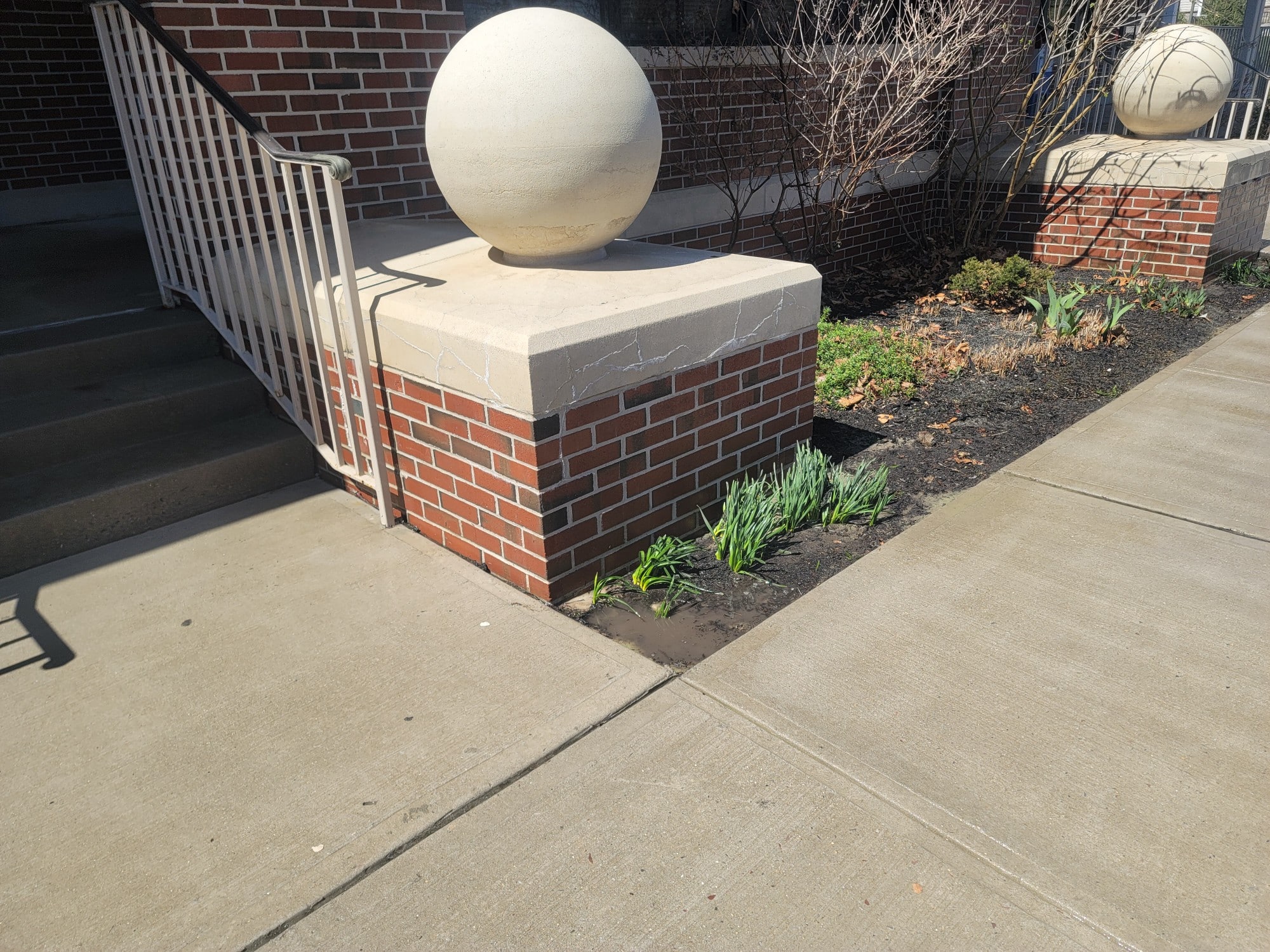 A brick structure with a spherical stone ornament on top stands next to a set of stairs. The adjacent garden has young green plants growing. The sidewalk in front of this commercial property is clean and well-maintained. A similar structure is visible further along the sidewalk.
