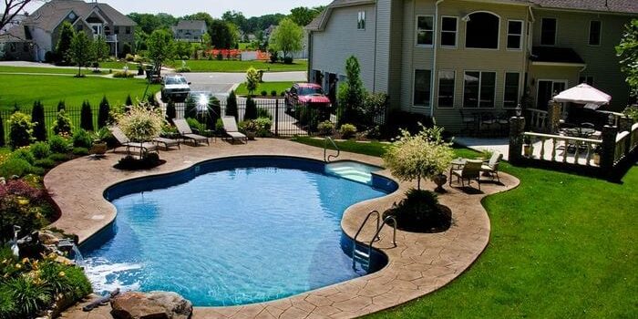 The backyard features a kidney-shaped swimming pool surrounded by a stone patio. Lounge chairs are placed around the pool, highlighting the pristine pool area cleaning. A well-manicured lawn and garden plants border the pool area, with a large house featuring an outdoor deck and a red car in the driveway in the background.
