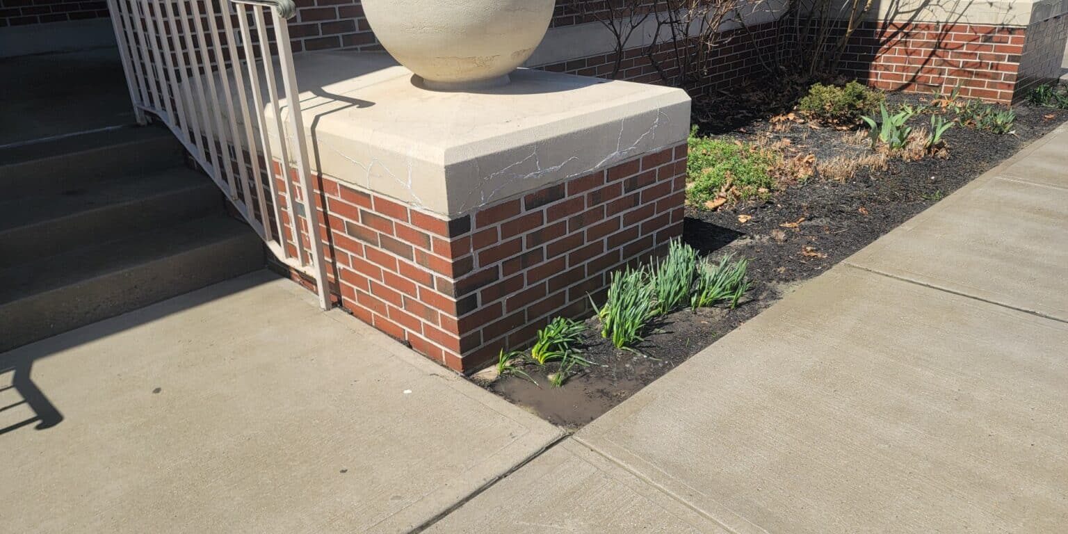 A brick structure with a spherical stone ornament on top stands next to a set of stairs. The adjacent garden has young green plants growing. The sidewalk in front of this commercial property is clean and well-maintained. A similar structure is visible further along the sidewalk.