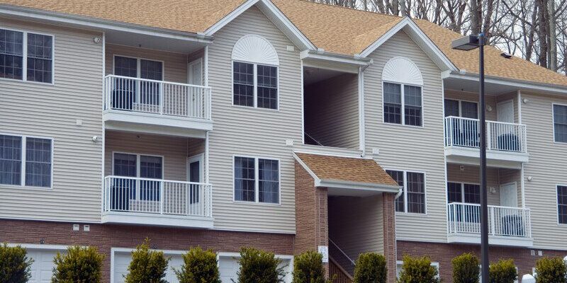 A two-story apartment building with beige siding and a brown roof features balconies with white railings. Bushes and small trees are planted in front of the building, and a streetlight is visible on the right side. The design allows for easy apartment cleaning, ensuring every unit remains pristine.