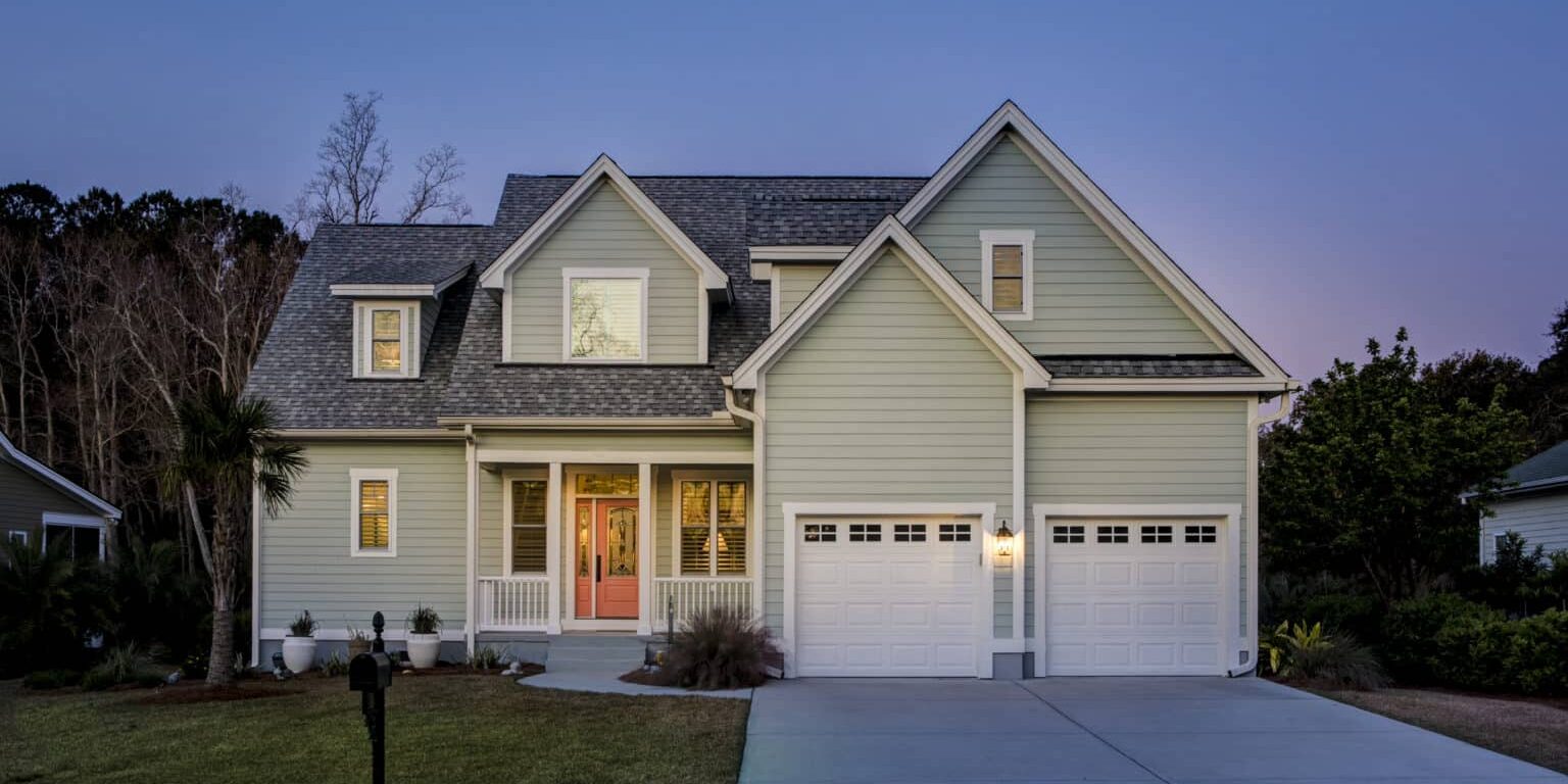 Two-story house with light green, clean siding and a two-car garage at dusk. The home boasts multiple windows, a well-maintained lawn, and significant curb appeal with its lit front porch featuring columns. A driveway leads to the garage doors, and trees are visible in the background.