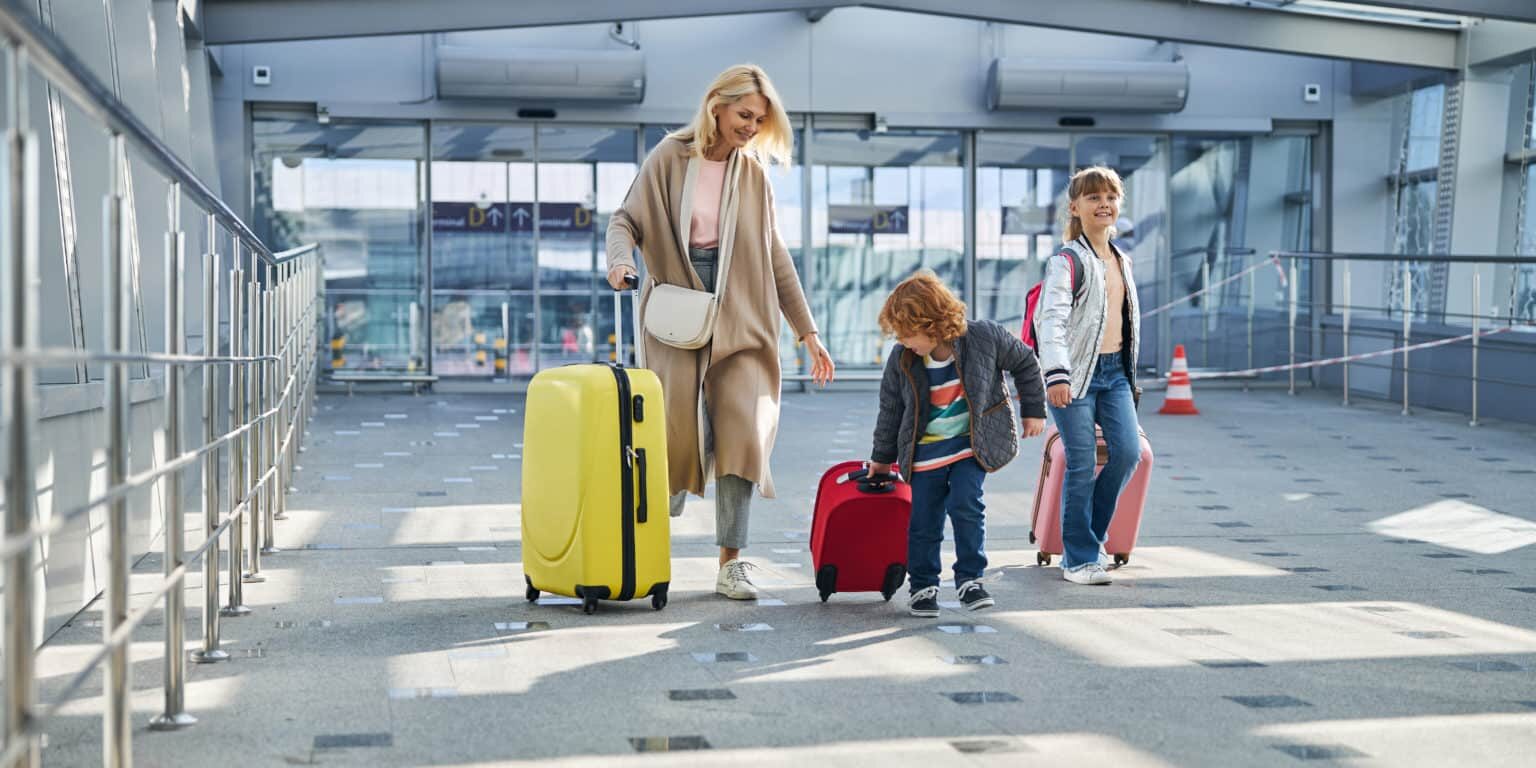 A woman and two children, ready for their summer vacation, walk through an airport with their luggage. The woman, in a long beige coat, carries a large yellow suitcase. The children, one with a red suitcase and the other with a pink backpack, follow closely. The background features modern architecture.