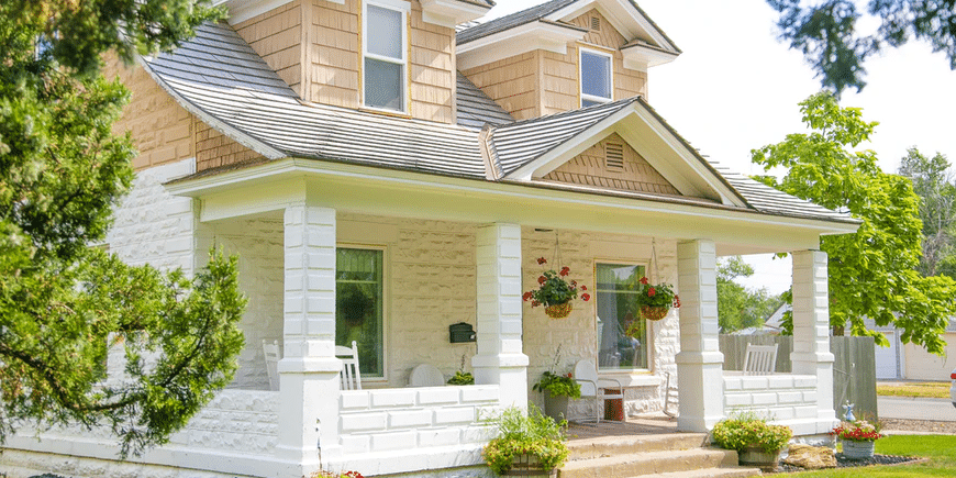 A charming white house with a cozy porch adorned with hanging flower baskets and potted plants. The home's exterior features dormer windows on the upper level and a well-maintained lawn, revived each spring. Tall trees frame the scene, adding to the peaceful, inviting atmosphere.