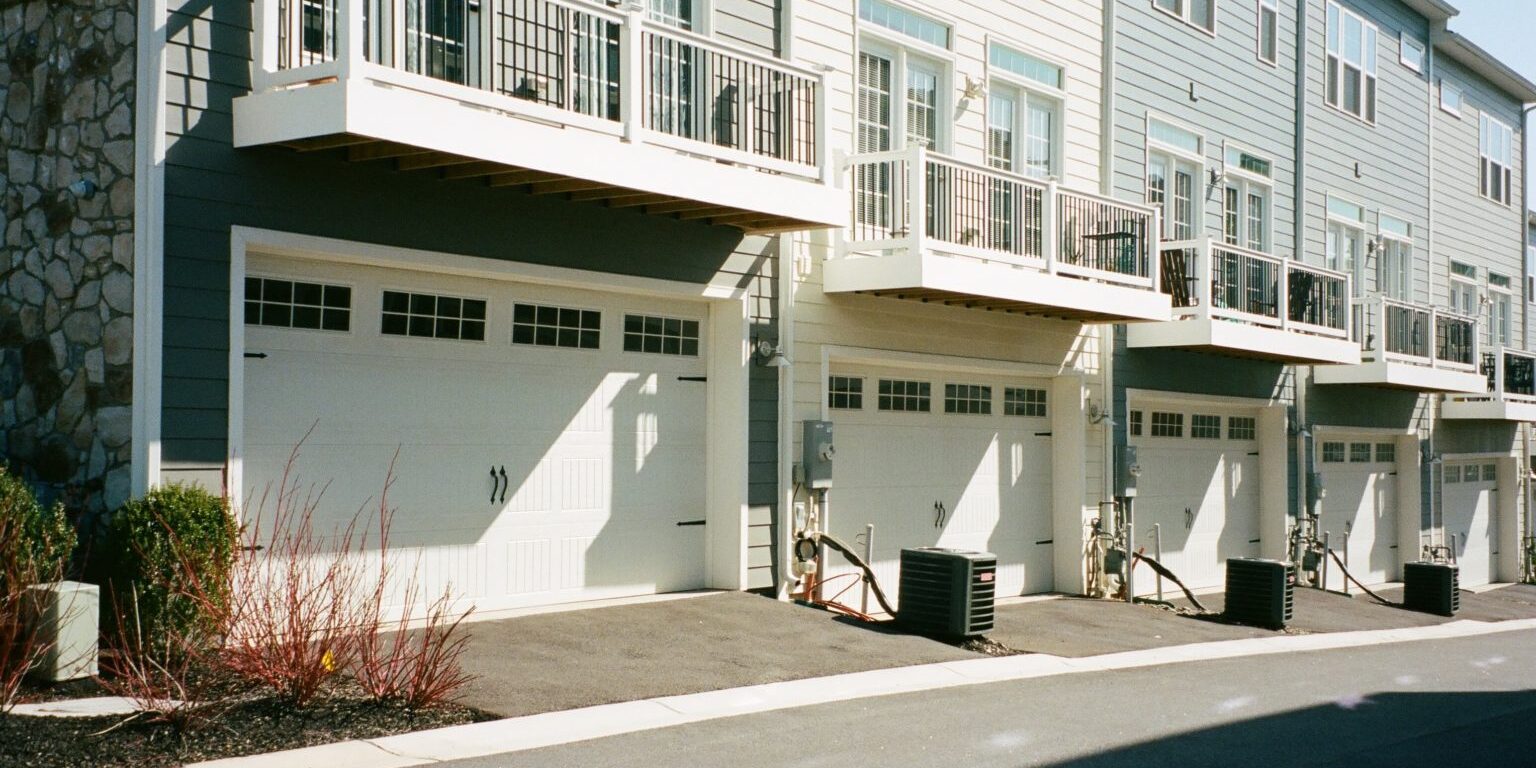 A row of modern townhomes with beige and gray exteriors. Each unit has a white garage door, balconies with railings above the garages, and air conditioning units beside the driveways. The street in front is clean and vacant, with minimal landscaping.