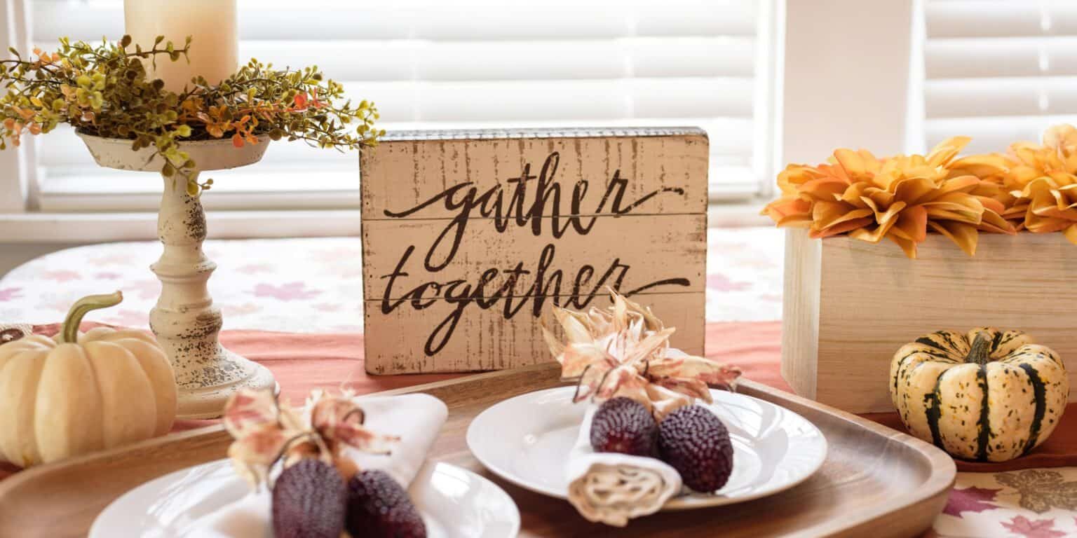 A cozy dining table setup for Thanksgiving featuring a "gather together" sign, decorative pumpkins, and dried corn on white plates. The table is adorned with a candle centerpiece and orange flowers, creating a warm, autumnal ambiance to help you prepare your home for the holiday season.